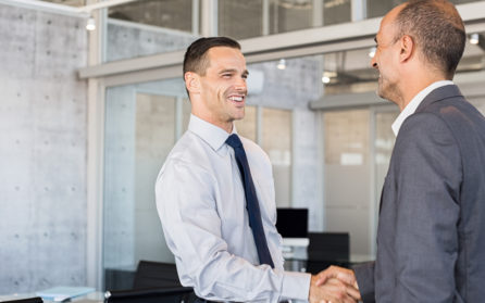 Two smiling businessmen shaking hands while standing in modern office boardroom. Happy business men shaking hands and looking at each other.